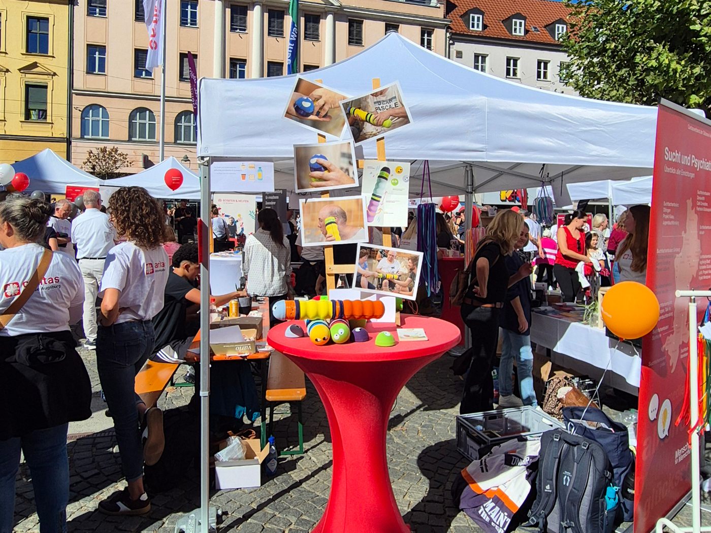 Unser Stand zwischen den weißen Zelten auf dem Moritzplatz: Auf einem roten Stehtisch liegen eine zusammengesetzte Maupe und einzelne bunte Teile, dahinter hängen Fotos von Menschen, die die Maupe in den Händen halten.