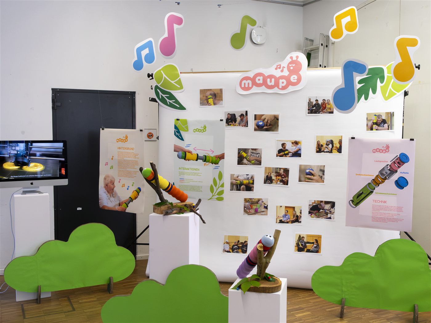 Unser Stand beim Sichtraum: An einer weißen Wand hängen viele Fotos und Plakate. Davor liegen zwei Maupen auf Ästen. Um sie herum stehen grüne Büsche aus Pappe.