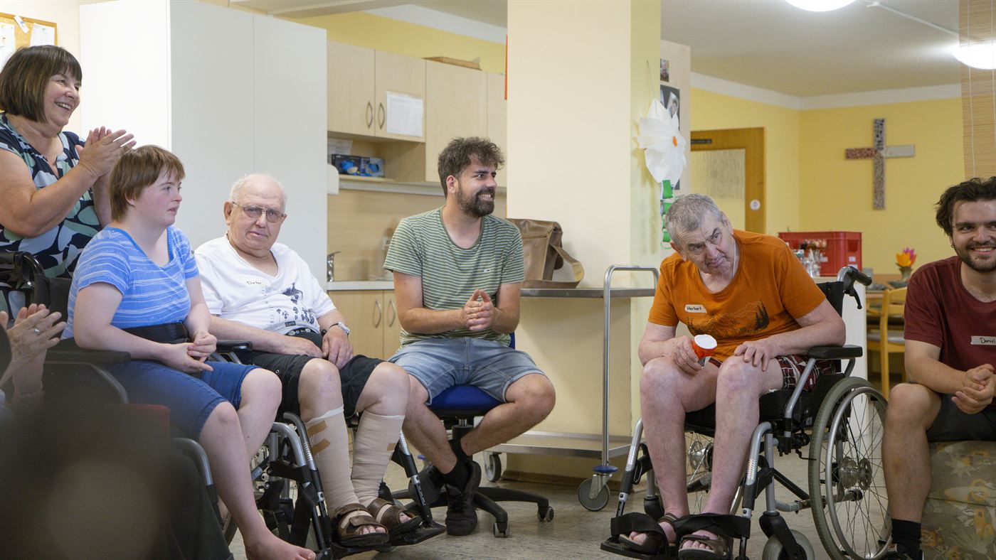 Eine Gruppe aus Bewohnerinnen, Bewohnern und Betreuenden sitzt im Kreis im Gemeinschaftsraum.