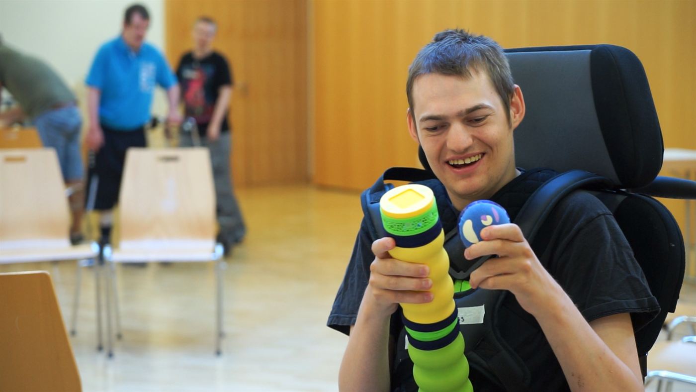 Ein junger Bewohner im Rollstuhl lacht und hält Maupe-Körper und Kopf in beiden Händen.